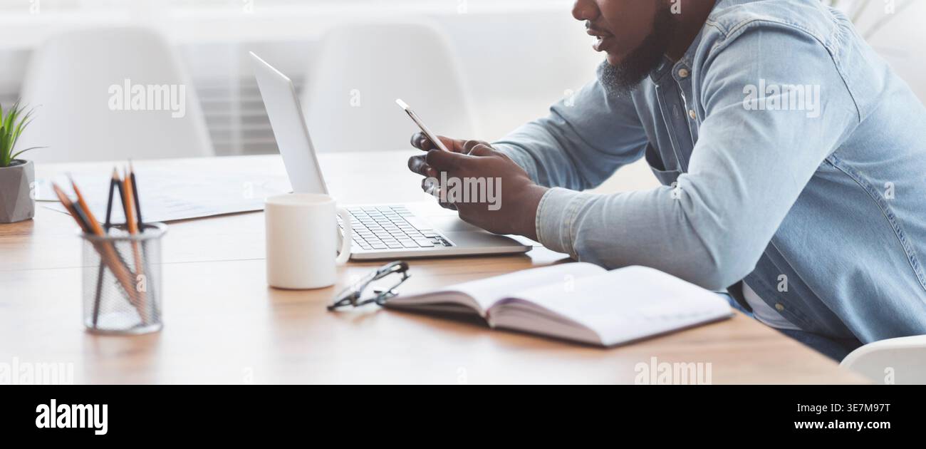 Black businessman using smartphone sitting at table with laptop in office Stock Photo