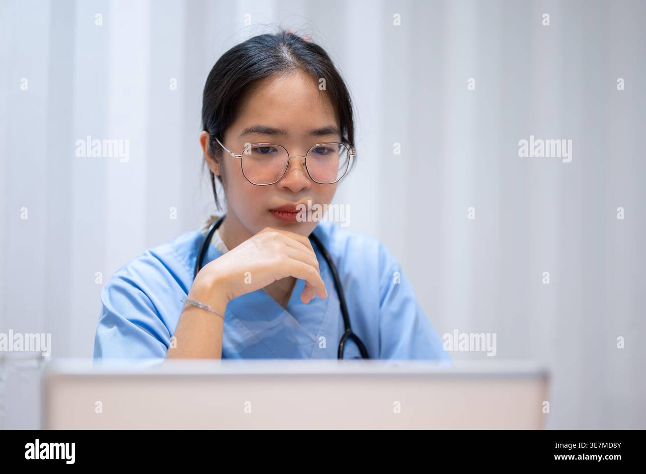 Asian female doctor in eyeglasses working on her laptop while sitting at table in hospital Stock Photo