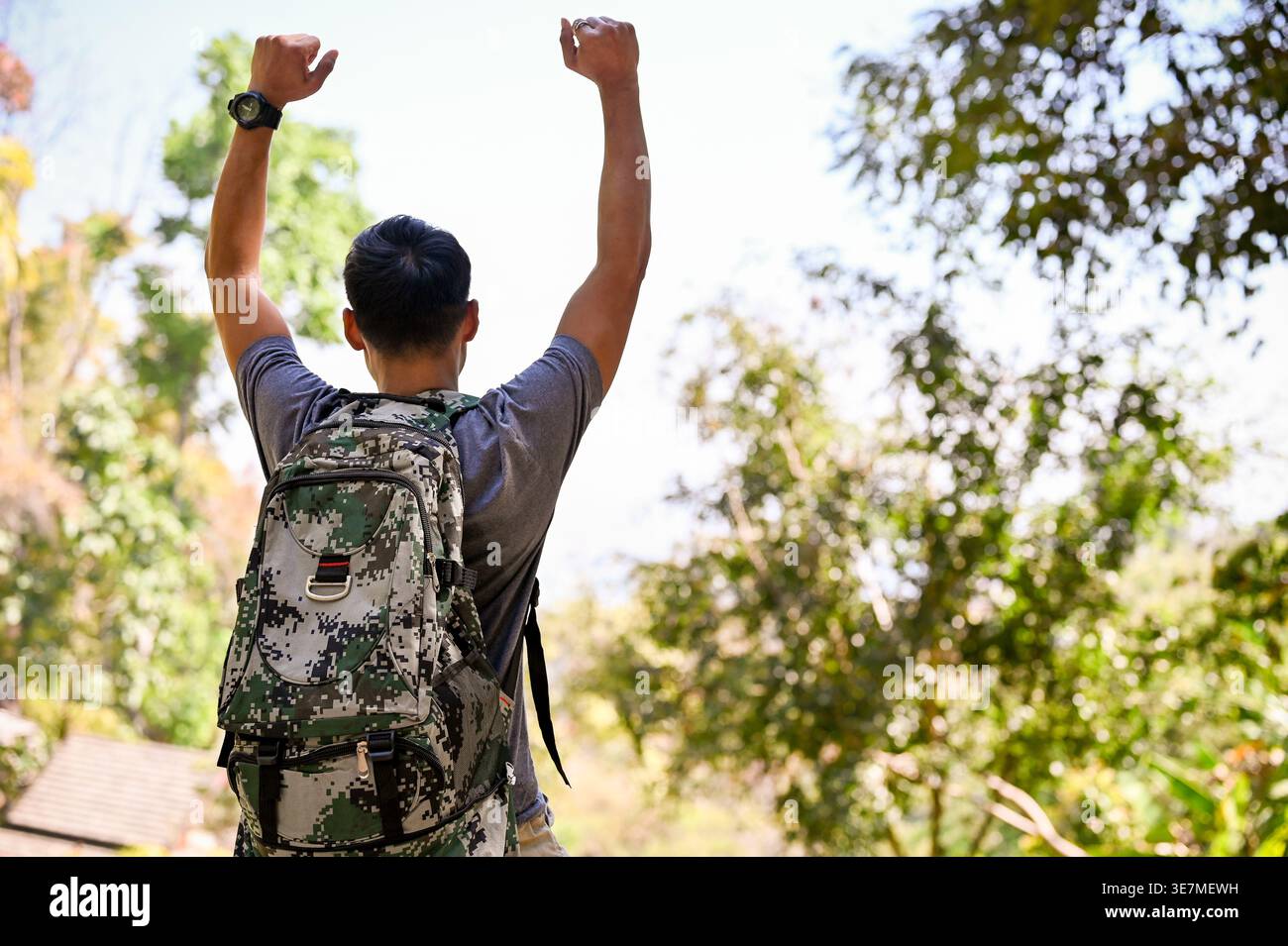 Asian male traveler raising arms while standing with backpack in forest on sunny day Stock Photo