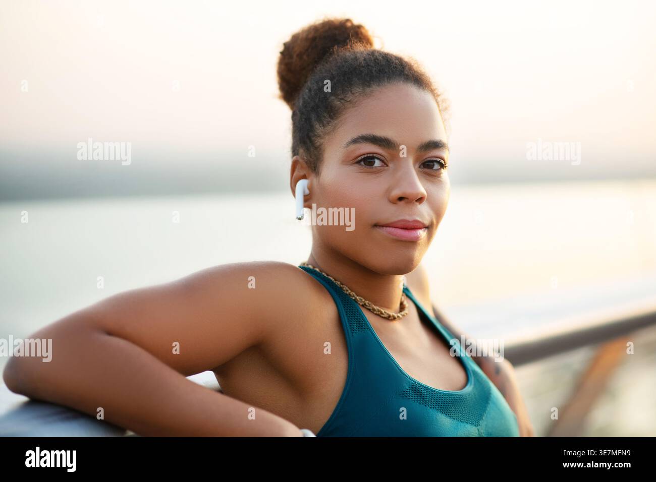 Smiling Mixed race woman in wireless earphones leaning arms on railing while standing on promenade during daytime Stock Photo