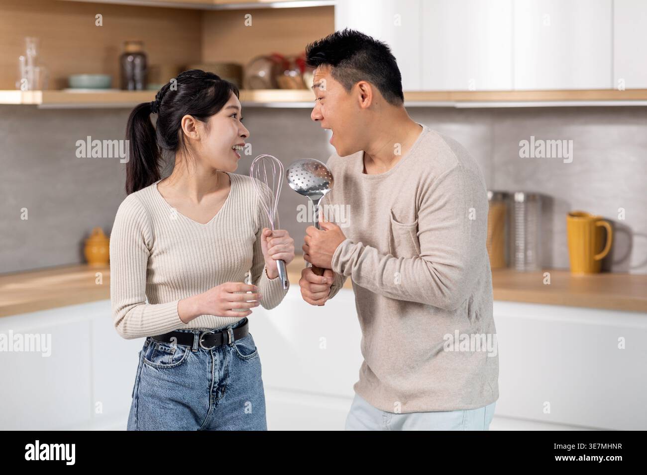 Cheerful Asian couple having fun singing using kitchen utensils as microphones while standing together at home Stock Photo