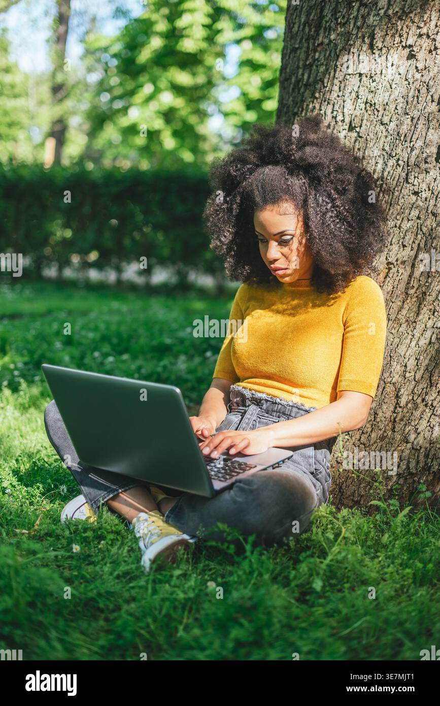 Hispanic female with curly hair working on laptop sitting on grass leaning on tree trunk in park during daytime Stock Photo