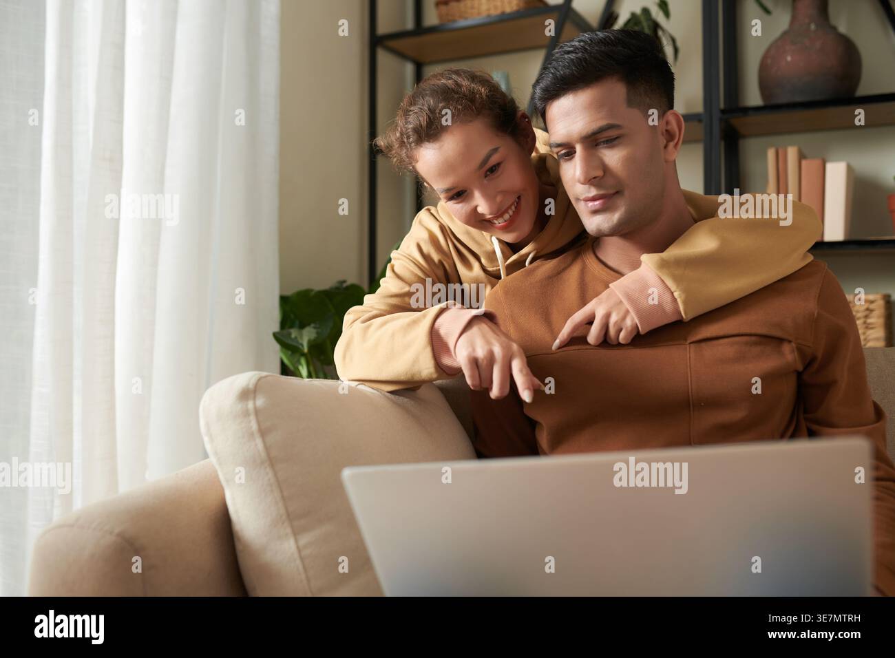 Smiling mixed race woman pointing finger and embracing Asian boyfriend from behind using laptop sitting on sofa in living room at home Stock Photo