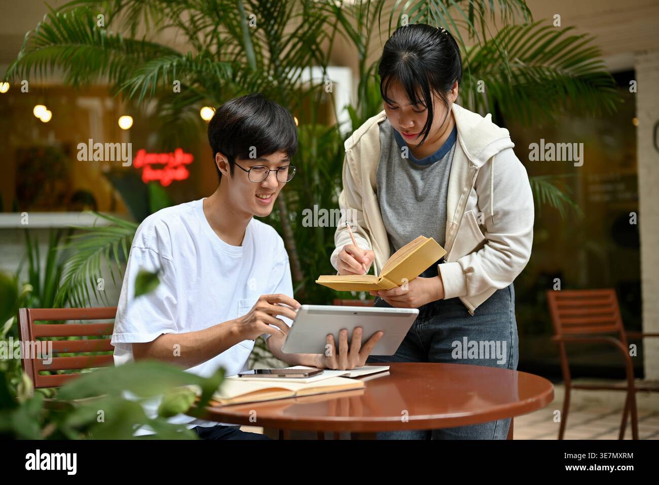 Smiling Asian man sitting at table using tablet with Asian female friend standing beside him and writing notes studying together in outdoor cafe Stock Photo