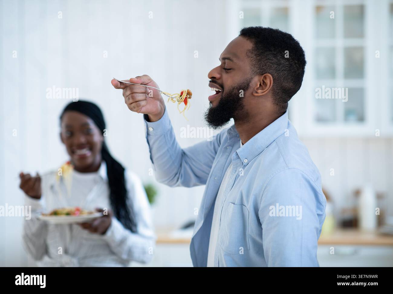 Black man eating spaghetti using fork standing in kitchen near wife at home Stock Photo