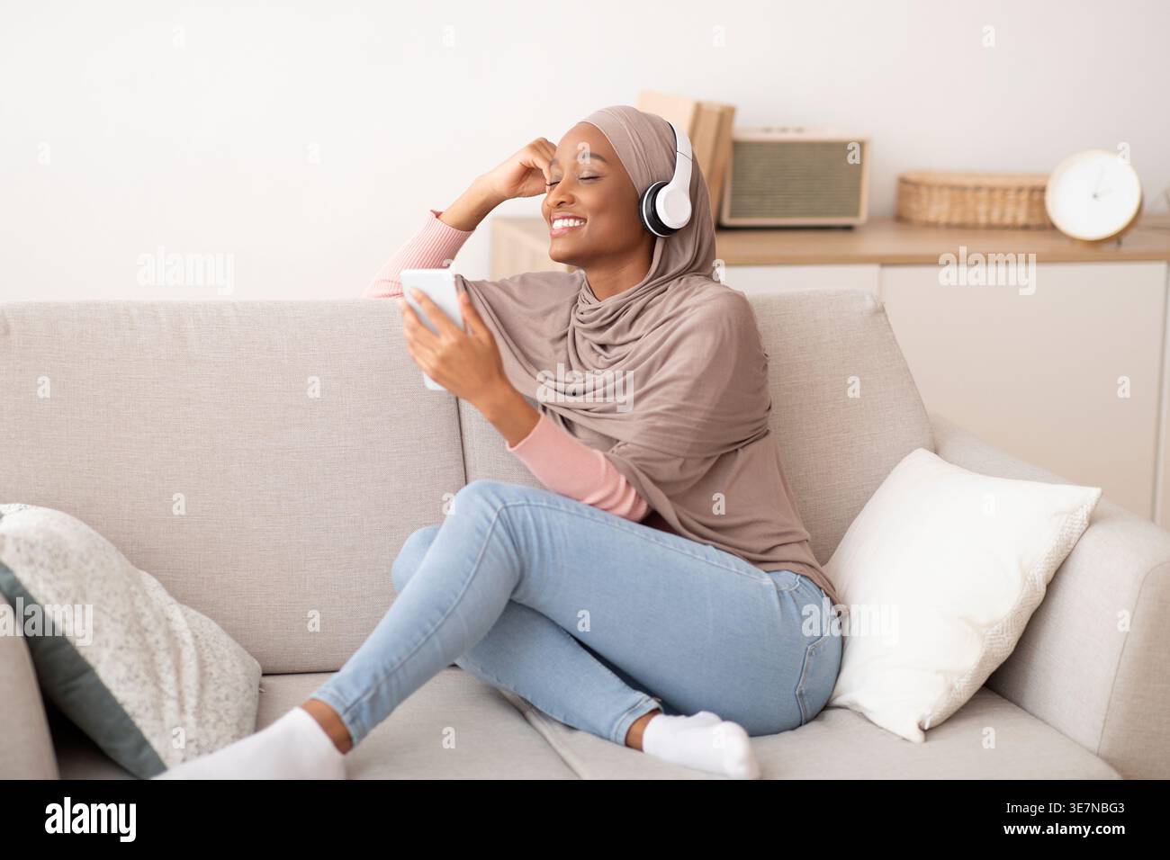 Joyful black Muslim woman in hijab and headphones listening to music on cellphone, closing her eyes in delight, at home. Islamic lady enjoying her fav Stock Photo