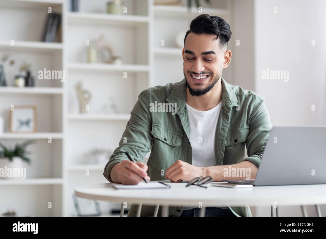 Smiling Arab man writing down notes sitting at table with laptop at home Stock Photo