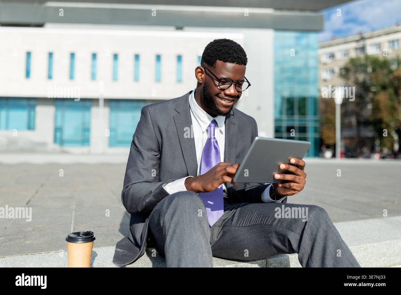 Black businessman in eyeglasses using tablet while sitting on stairs beside takeaway coffee cup outside the office building on sunny day Stock Photo