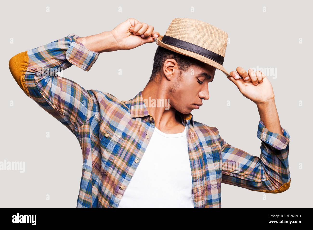 Black man with closed eyes tipping straw hat while standing against light gray background Stock Photo