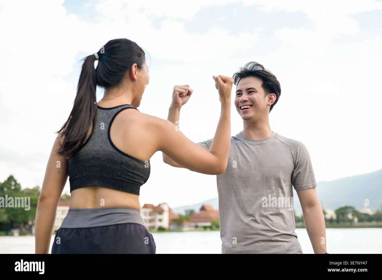 Cheerful Thai couple with raised arms and closed fists celebrating success after reaching their running target while standing together near lake in... Stock Photo