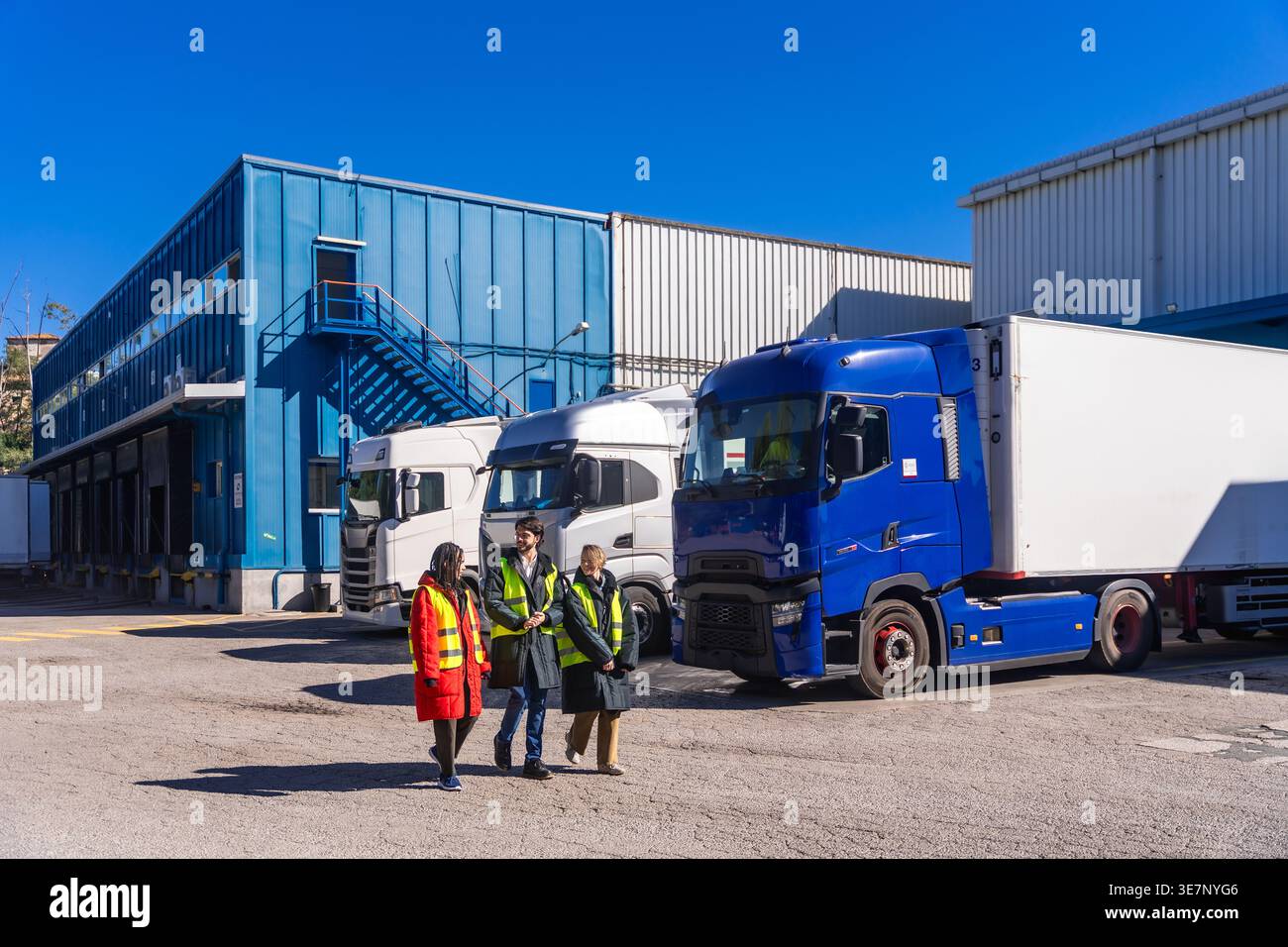 Diverse group of engineers wearing hard hats and safety vests walking together near transport trucks at cold storage warehouse on sunny day Stock Photo