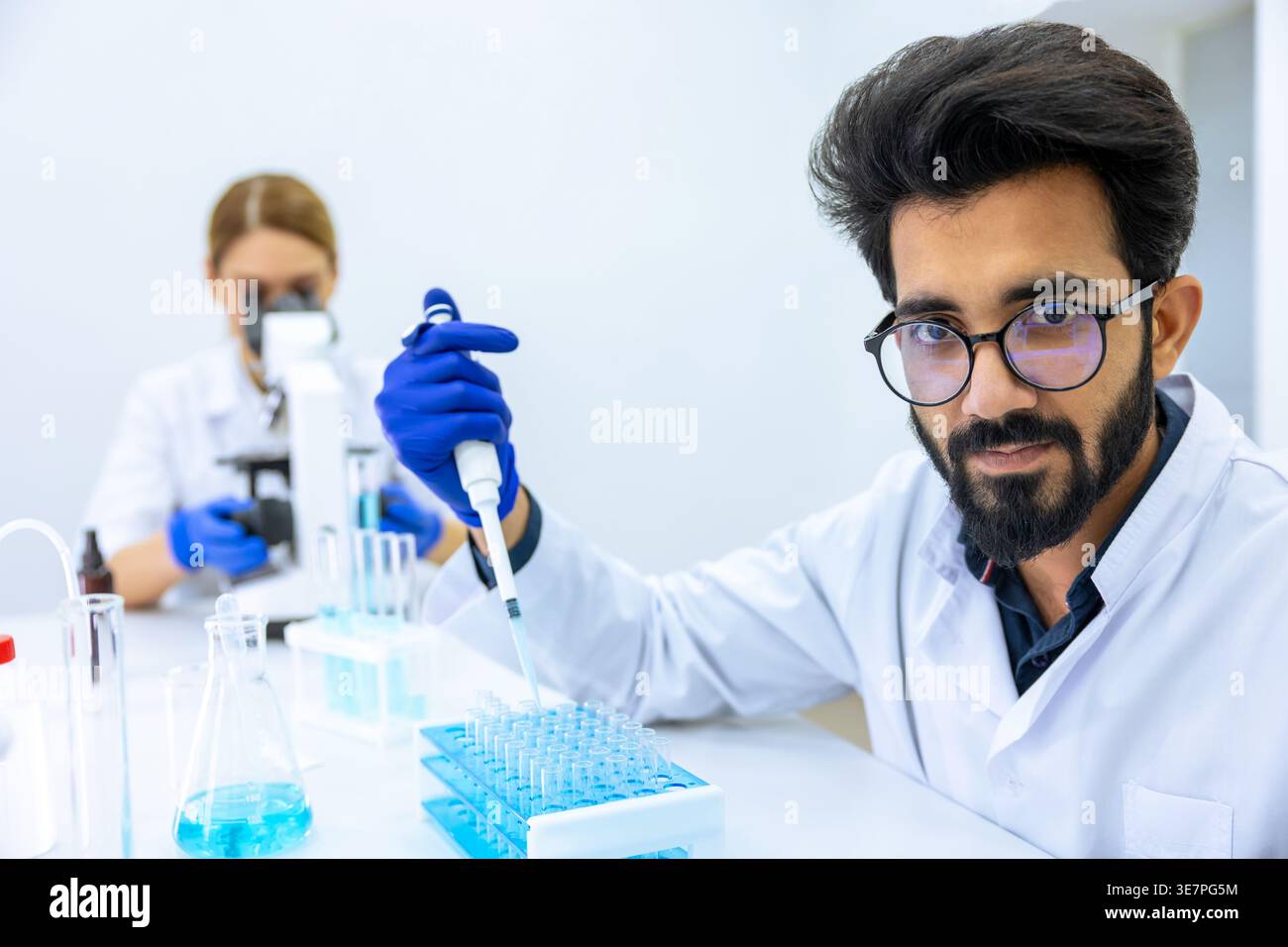 Indian male scientist working with chemical liquids in flask near caucasian female colleague using microscope in laboratory Stock Photo