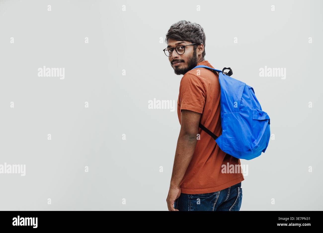 Serious Indian man in orange shirt with eyeglasses and blue backpack standing while holding notebook against white background in studio Stock Photo