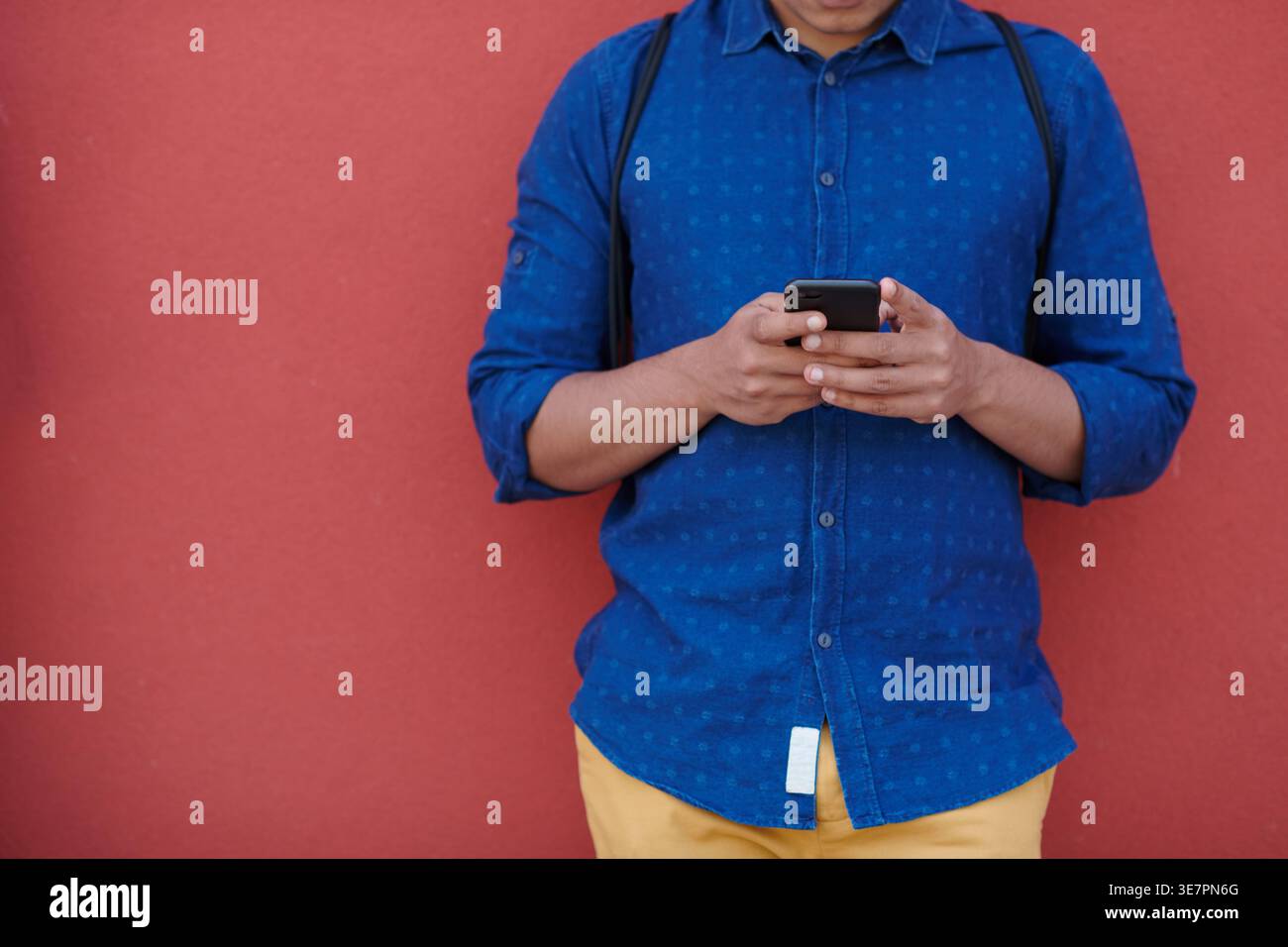 Middle Eastern man using smartphone leaning against red wall outdoors Stock Photo