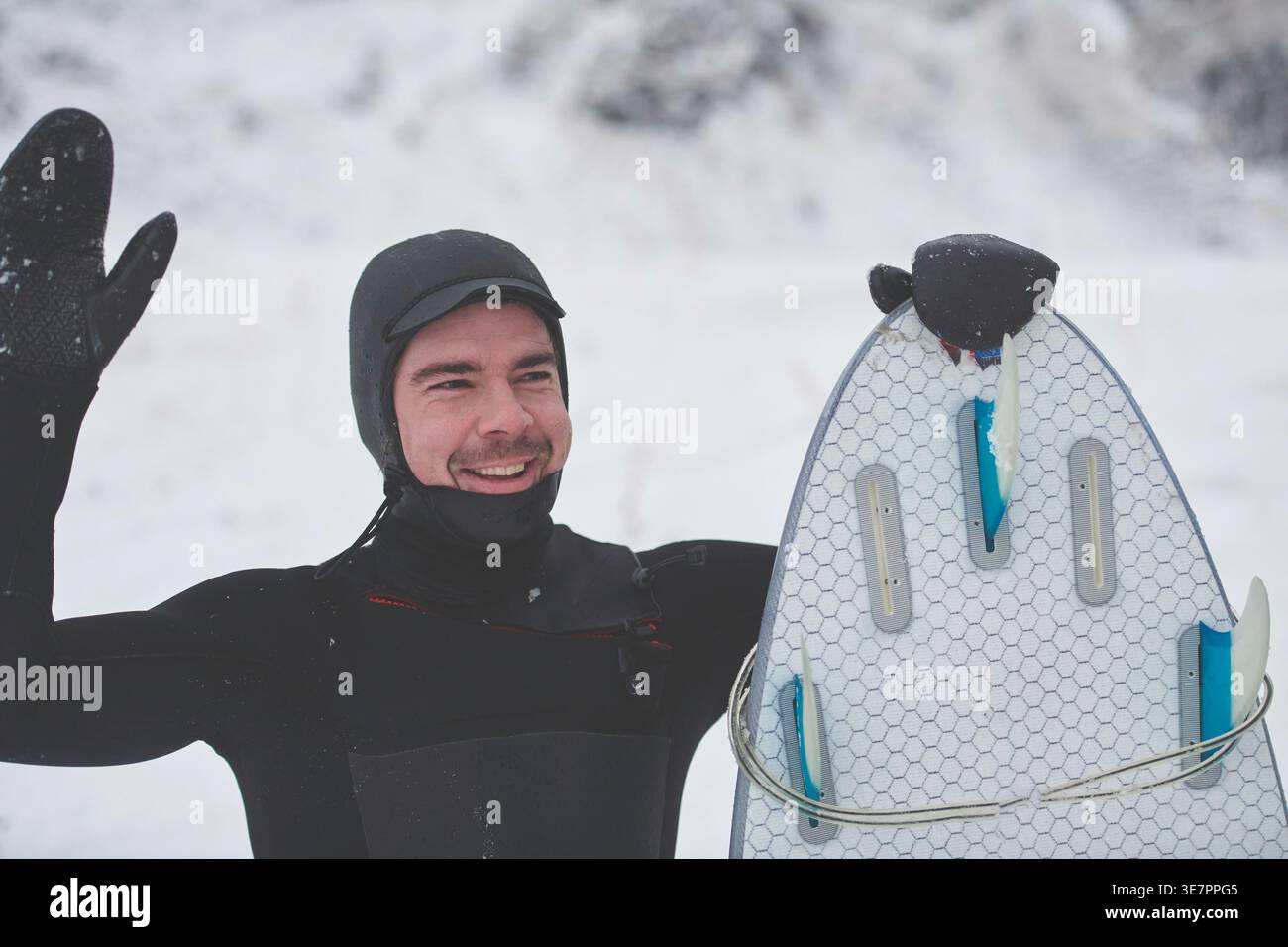 Smiling Mixed race man in surfing suit holding surf board and looking away while standing on snowy mountain during winter Stock Photo