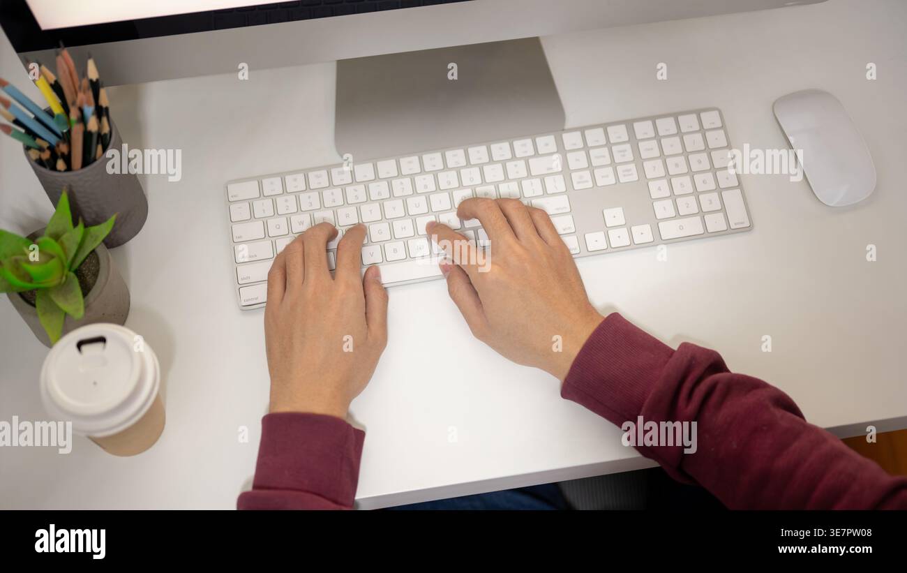 Man wearing maroon hoodie using computer while sitting at table in office Stock Photo