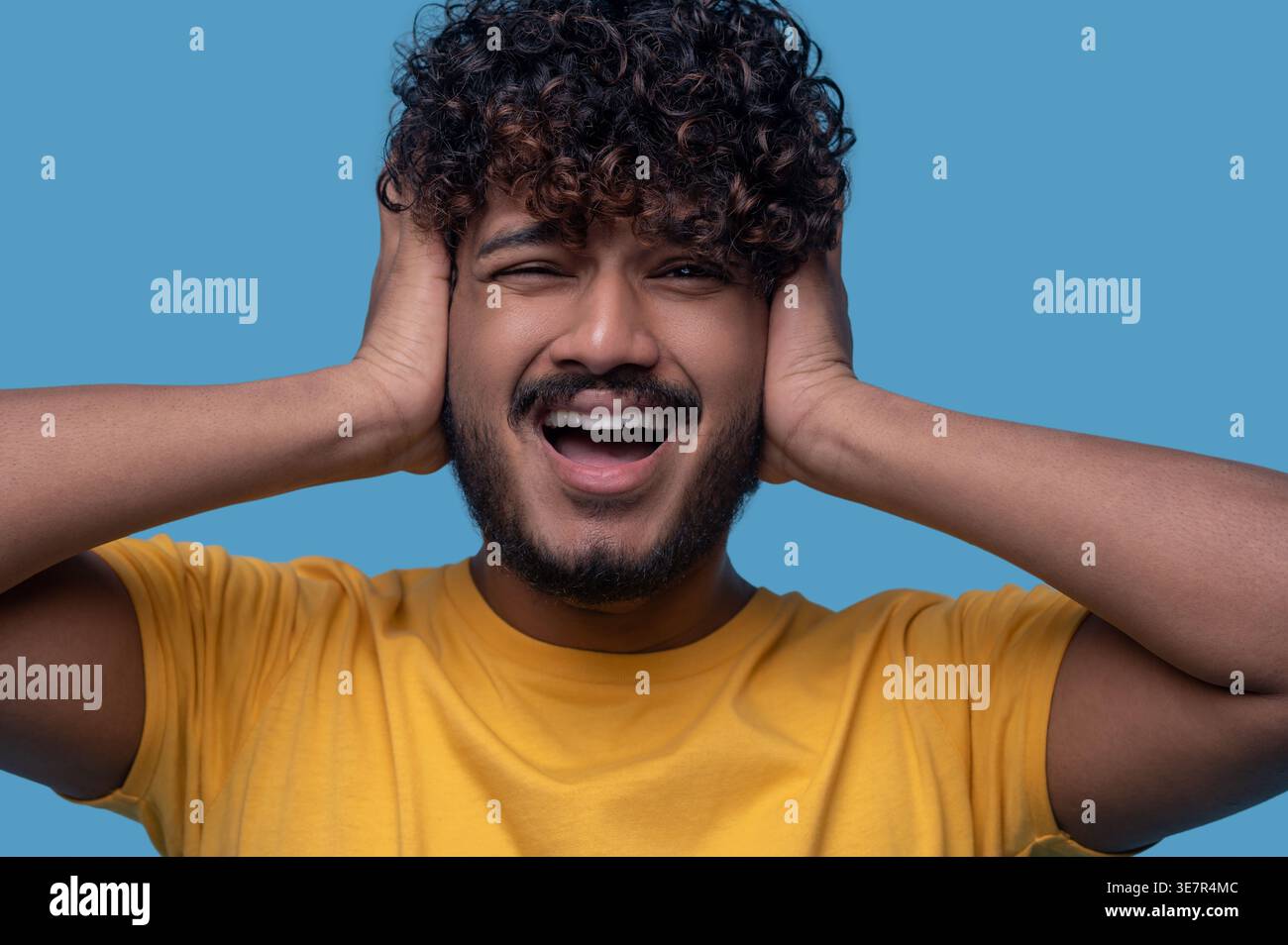 Indian man with curly hair standing with hands covering ears and mouth opened against blue background Stock Photo