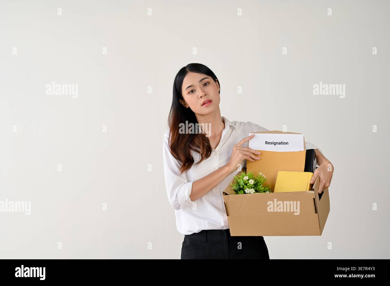 A tired and despairing young Asian businesswoman or female office worker holds a cardboard box with her stuff and a resignation letter, standing over Stock Photo