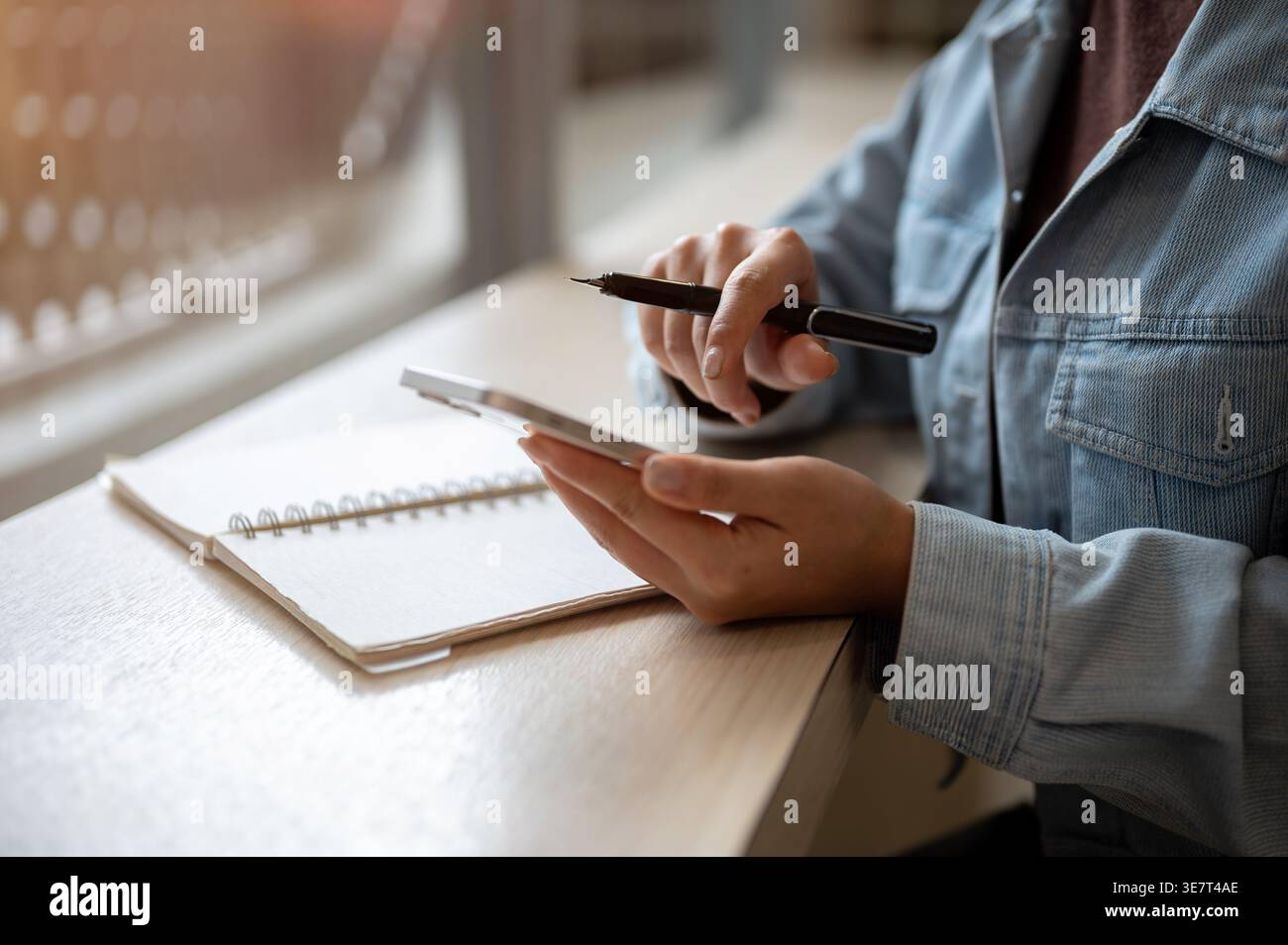 Cropped image of Thai woman in denim jacket browsing on smartphone and holding stylus pen while sitting at table with notebook indoors Stock Photo