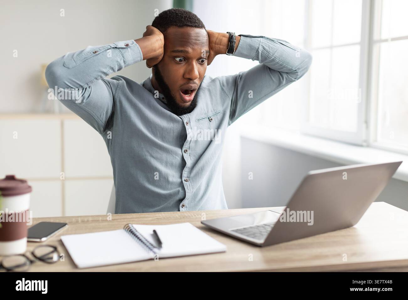 Shocked Black businessman touching his head and looking at laptop screen while sitting at table with smartphone, eyeglasses, disposable coffee cup,... Stock Photo