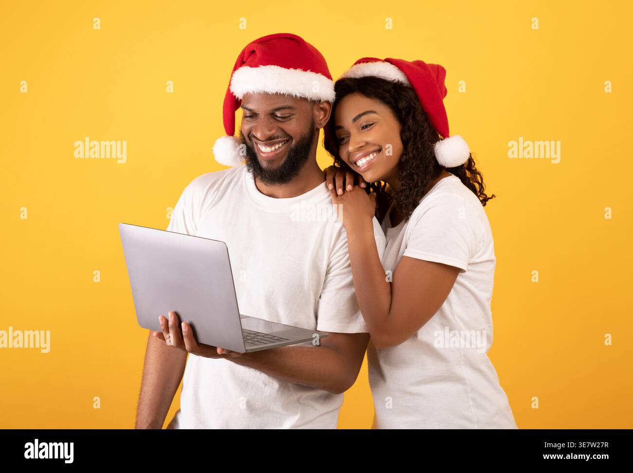 Smiling Black couple wearing Santa hats holding and looking at laptop standing against yellow background in studio Stock Photo
