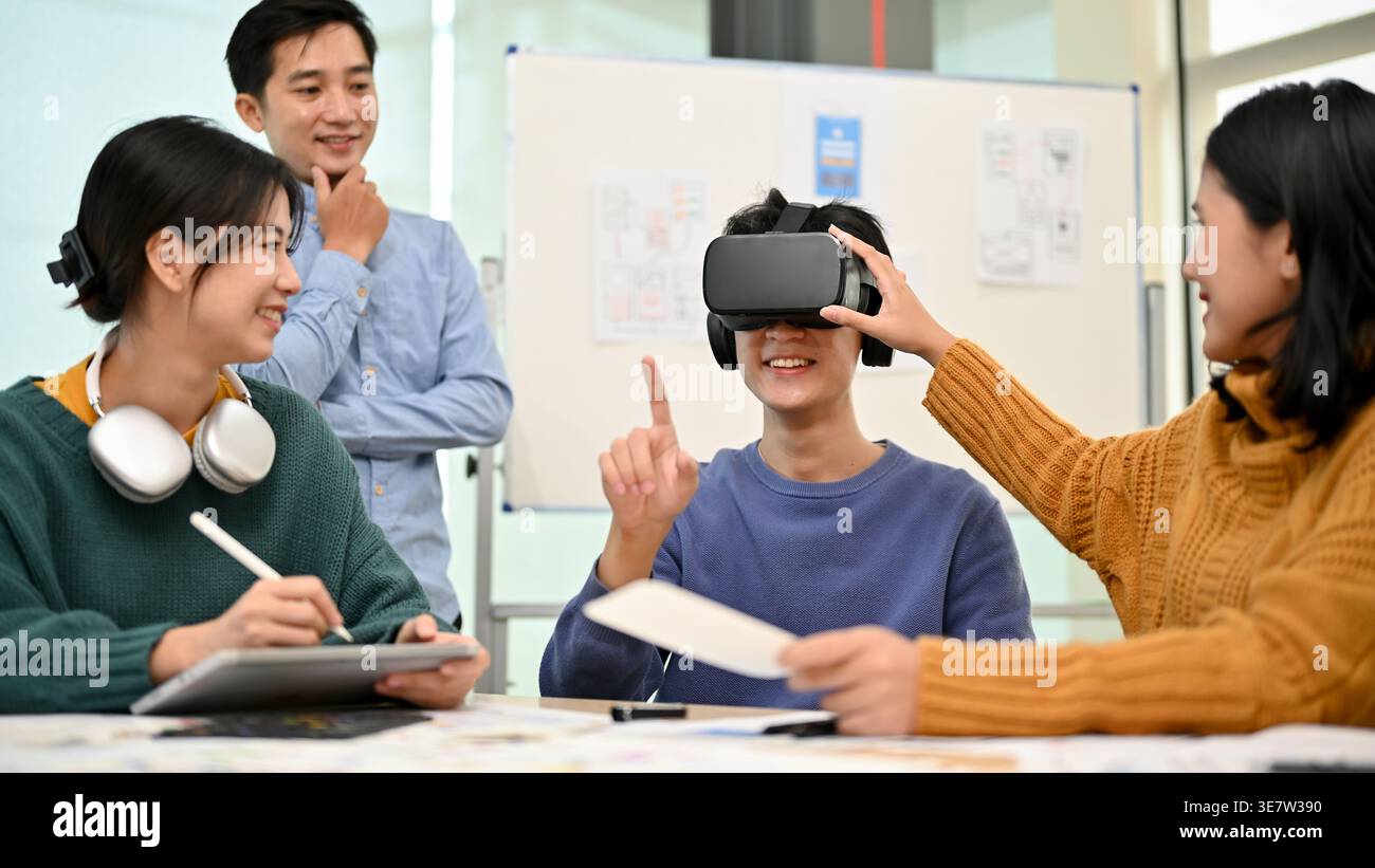 Smiling Asian male developer wearing VR headset sitting at table and testing newly developed VR game with colleagues in office Stock Photo