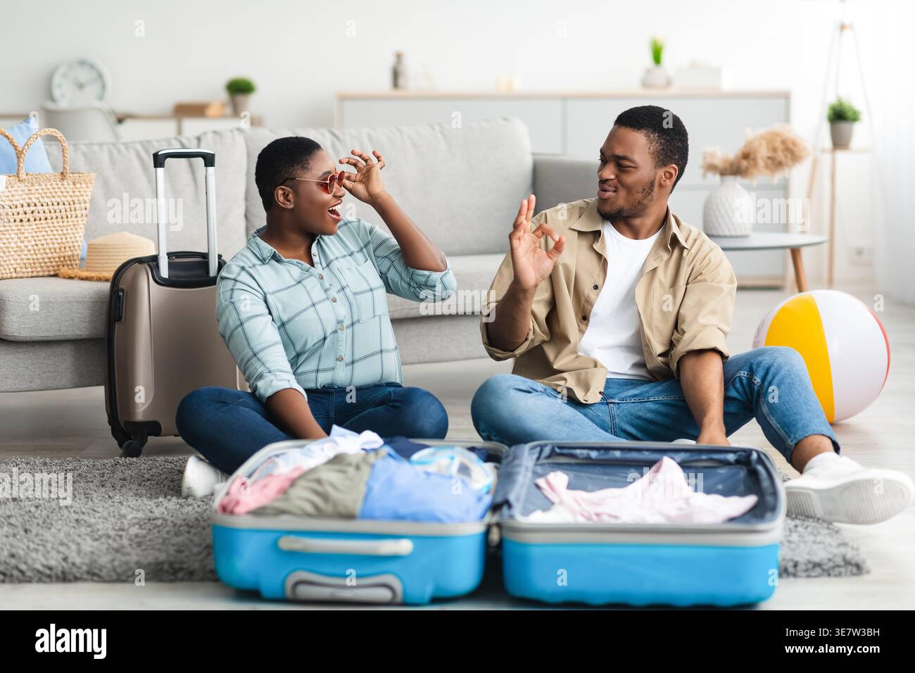 Cheerful Black man gesturing approving wifeâ€™s sunglasses while sitting on floor with packed suitcase in living room at home Stock Photo