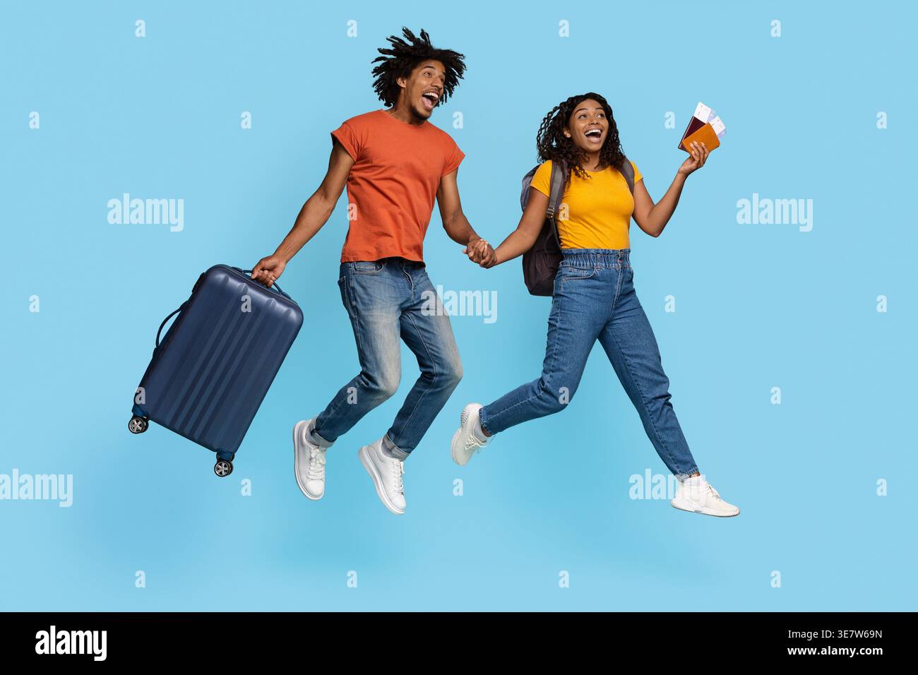 Cheerful Black man with open mouth carrying suitcase holding hands with girlfriend holding passports and tickets while jumping against blue backgro... Stock Photo