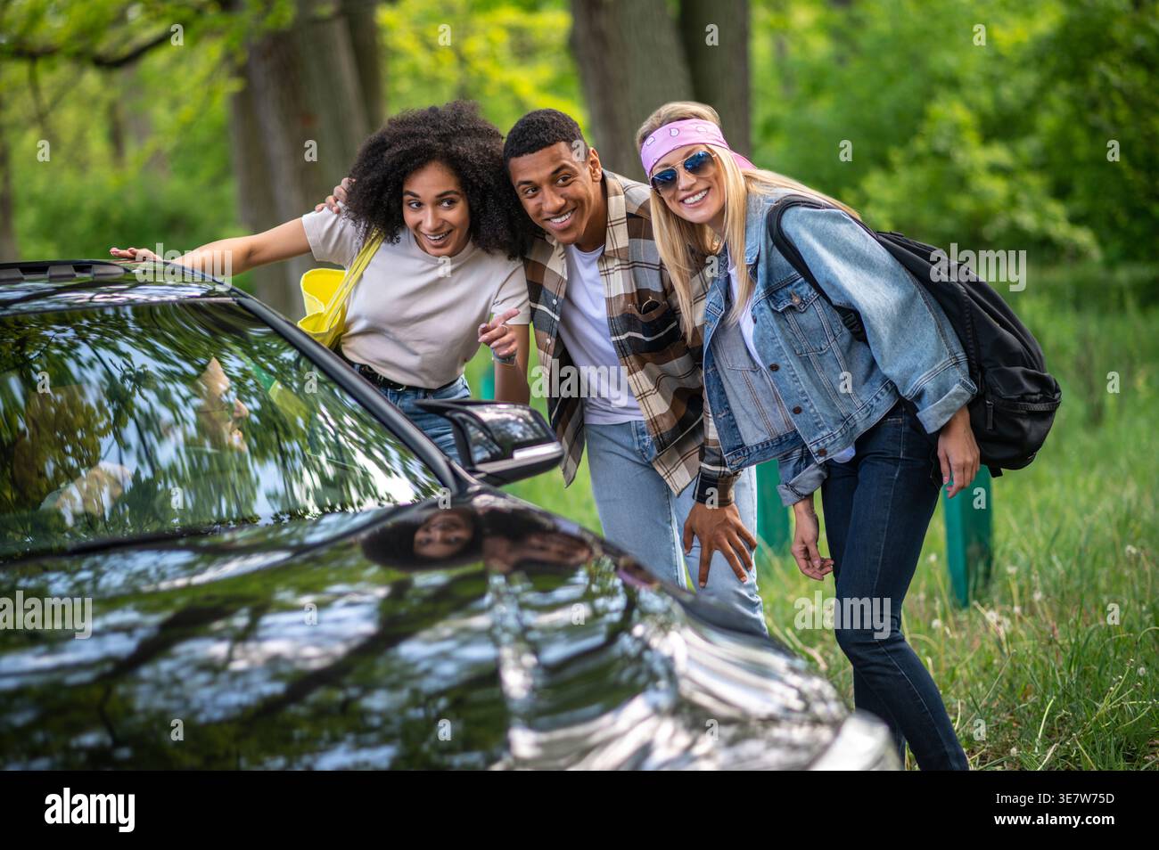 Smiling Black woman with male and female friends talking to the driver of black car pointing direction while standing on forest road on sunny day Stock Photo