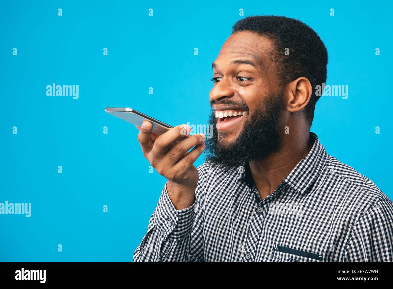 Black man recording voice message on smartphone while standing against blue background in studio Stock Photo