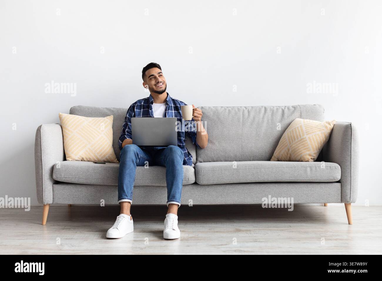 Middle Eastern man having coffee while using laptop sitting on sofa in living room at home Stock Photo