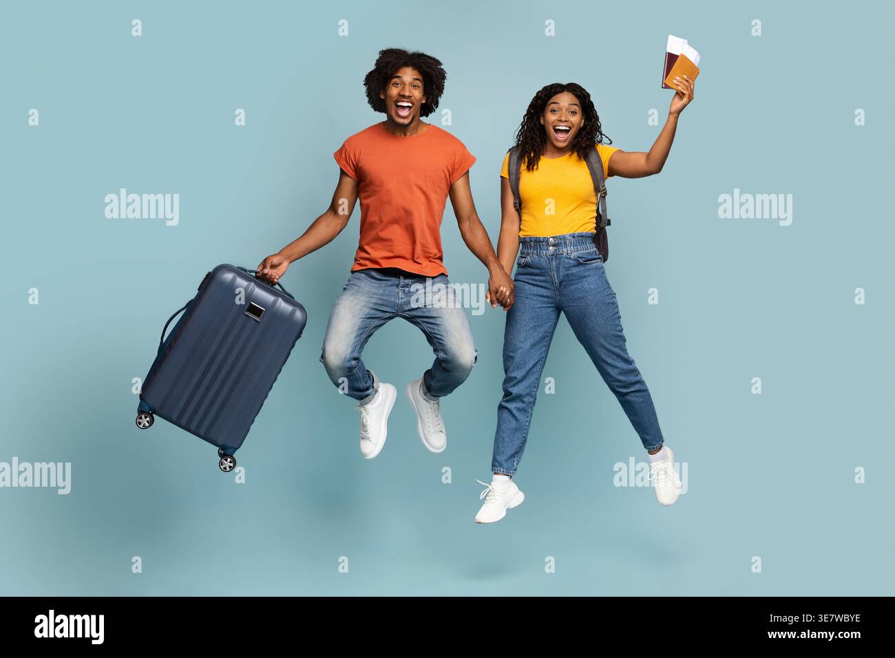 Cheerful Black man with open mouth carrying suitcase holding hands with girlfriend holding passports and tickets while jumping against blue backgro... Stock Photo