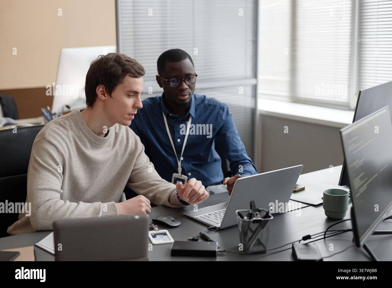 Black and white male IT specialists working on laptop together sitting at desk in office Stock Photo