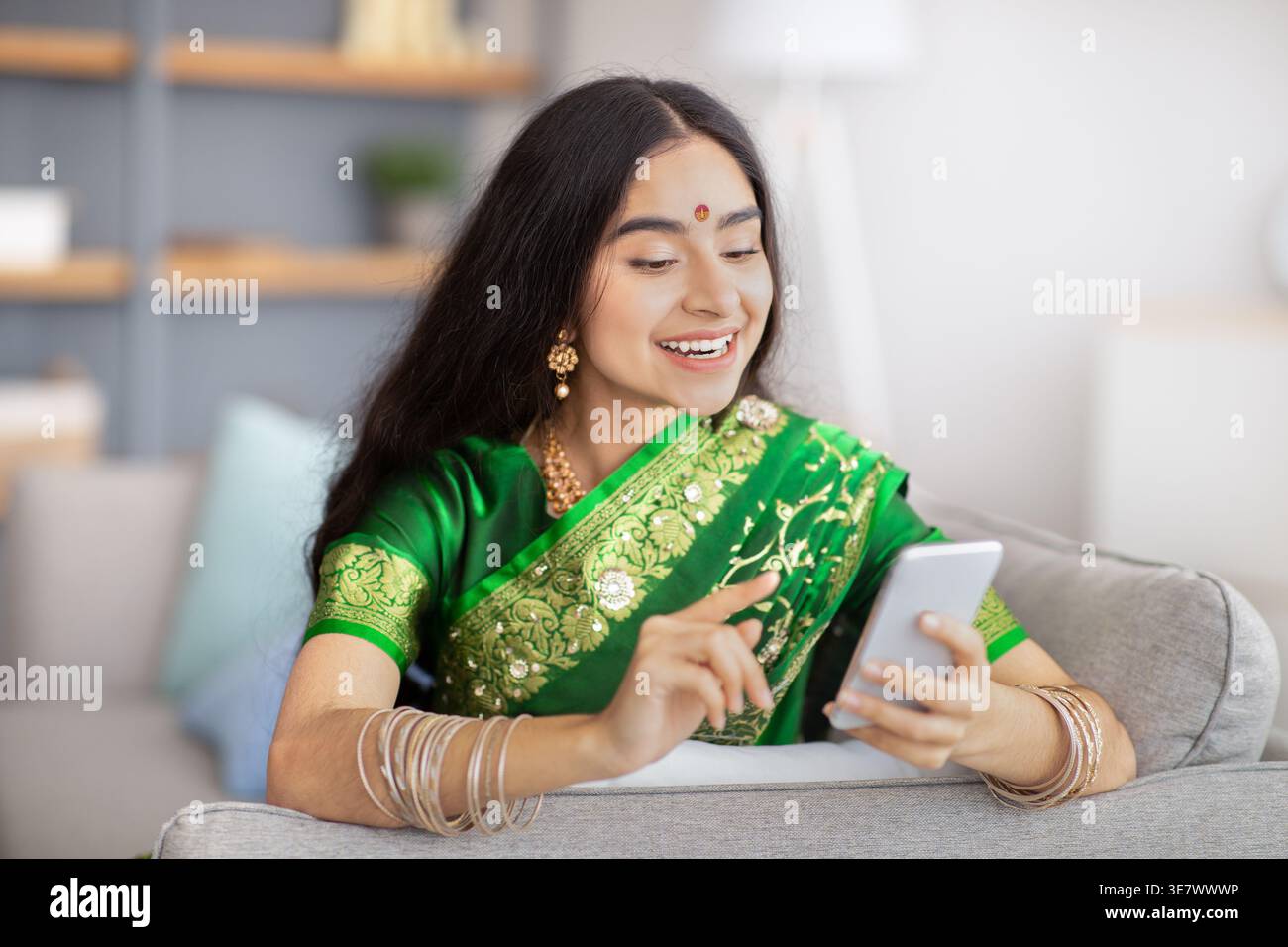 Smiling Indian female in traditional sari dress using smartphone sitting on sofa at home Stock Photo