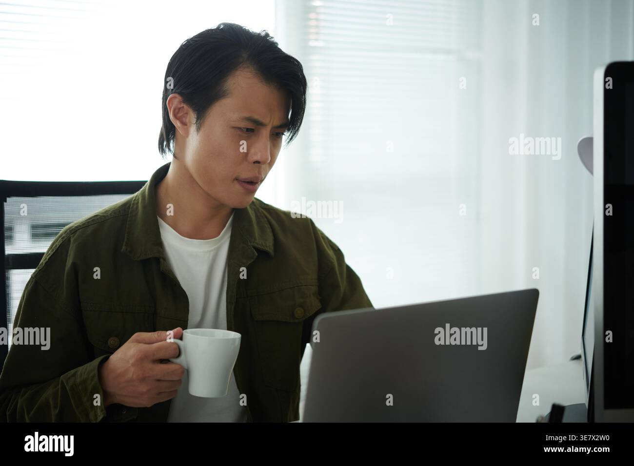 Asian male programmer drinking coffee and using laptop while sitting at table in office Stock Photo