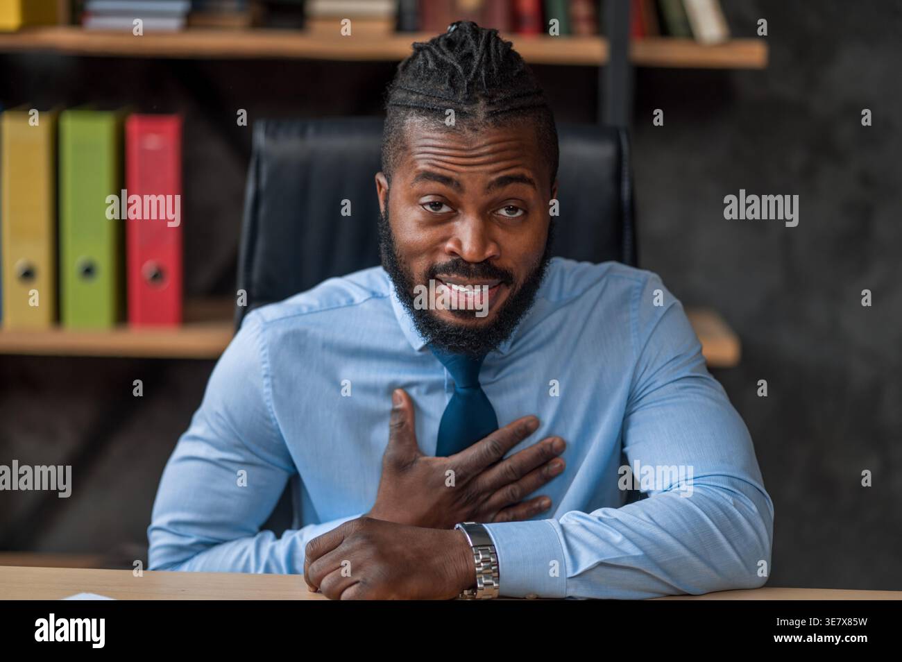 Black businessman with hand on chest sitting at table in office Stock Photo