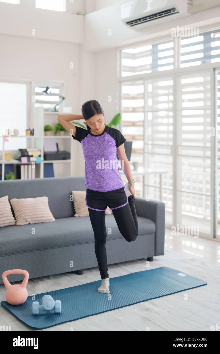 Young Asian woman stretches leg during indoor fitness session. Engaging in home workout for health strength wellness. Focused adult athlete. Stock Photo
