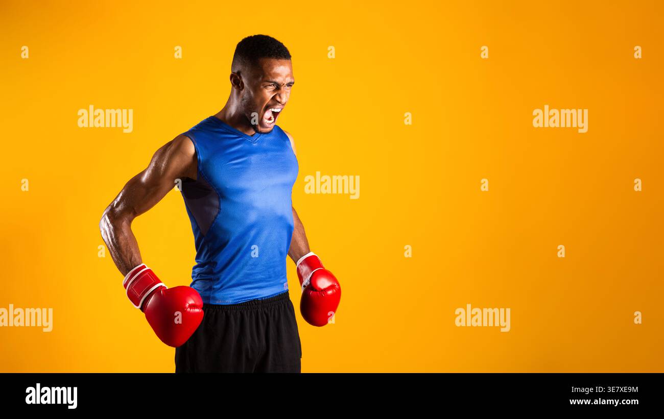 Black man wearing boxing gloves looking away while standing against yellow background in studio Stock Photo