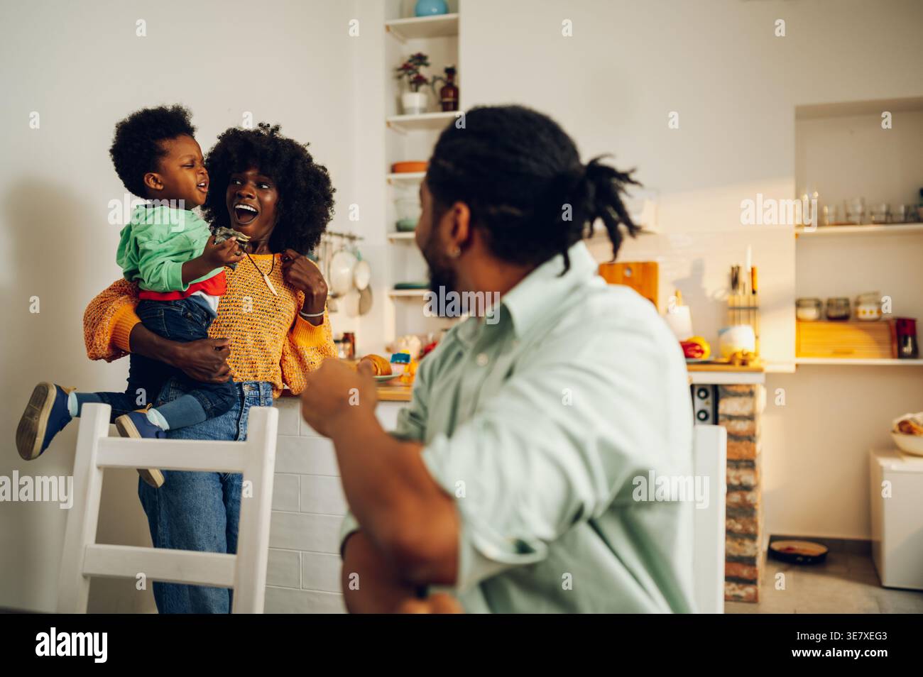 Cheerful Black mother with afro holding her son standing in kitchen near husband sitting at table and looking at them at home Stock Photo