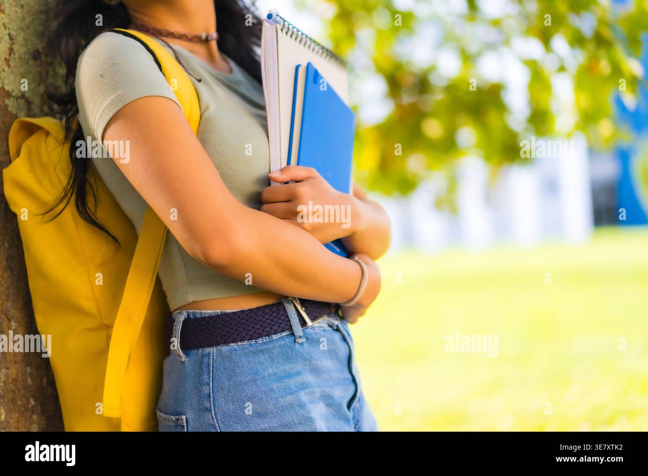 Asian female student with backpack holding books leaning against tree trunk in university during daytime Stock Photo