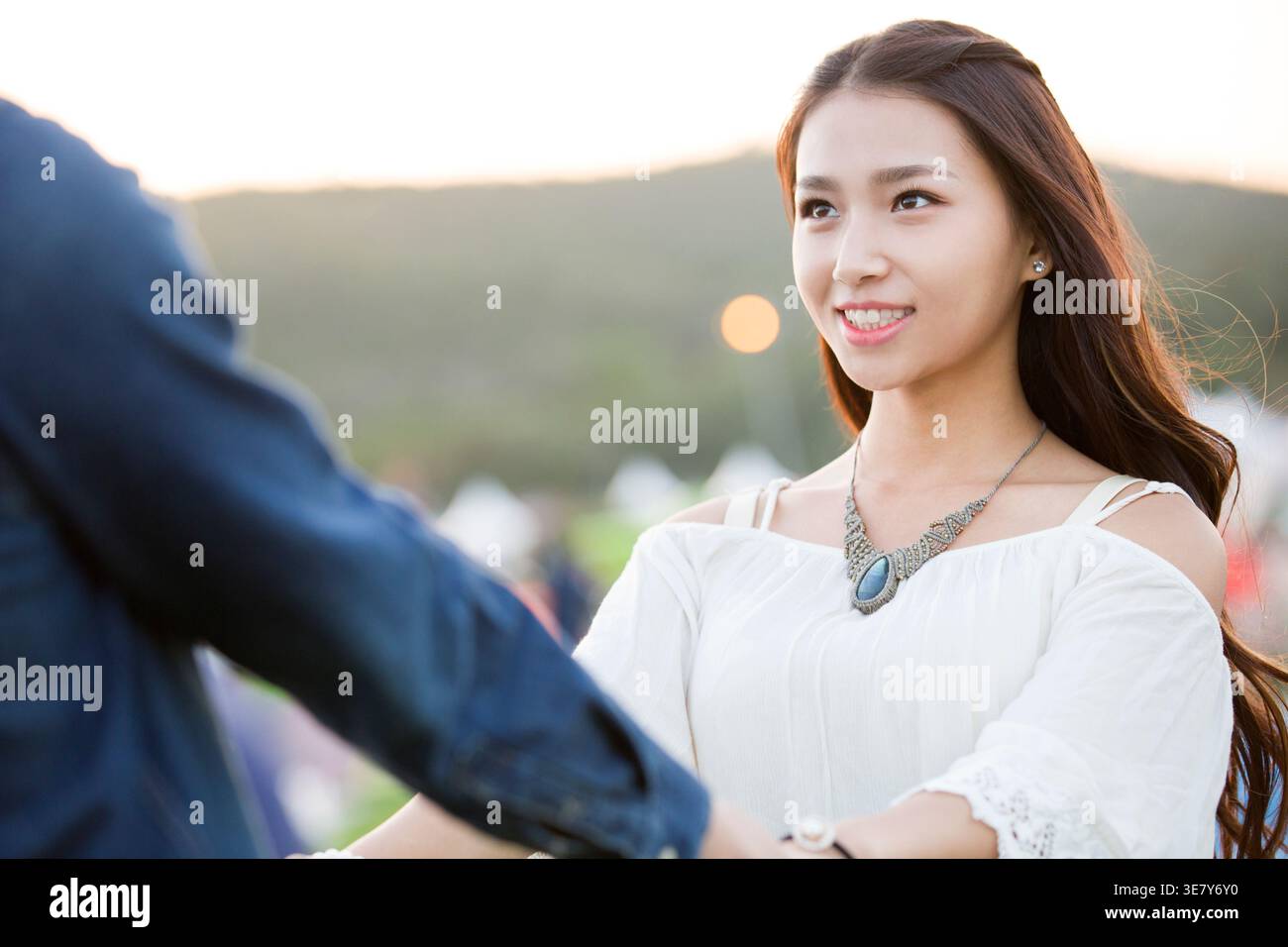 Smiling Chinese woman holding hands with boyfriend having fun while standing on concert ground with tents at music festival Stock Photo