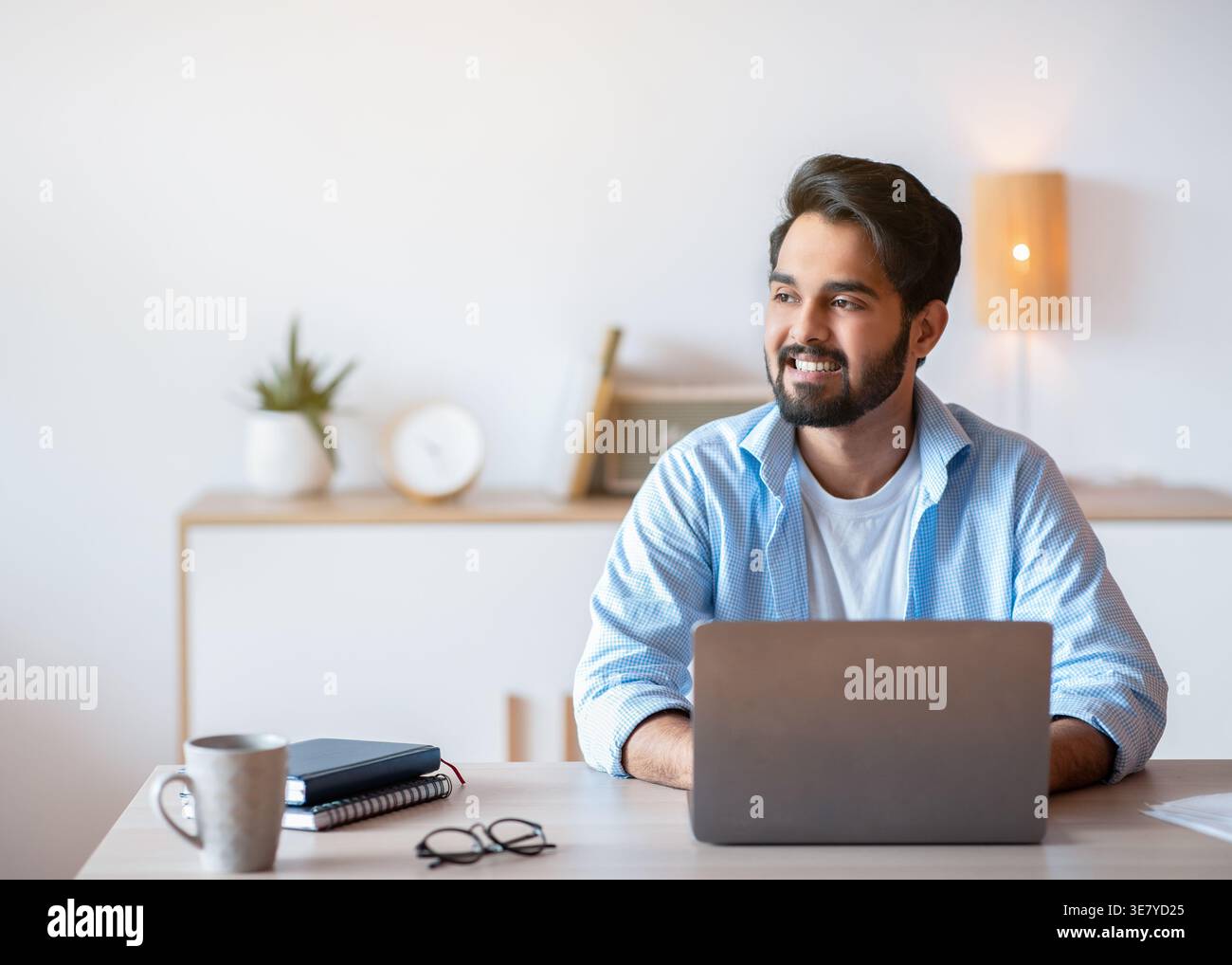 Smiling Middle Eastern man looking away working on laptop sitting at table at home office Stock Photo