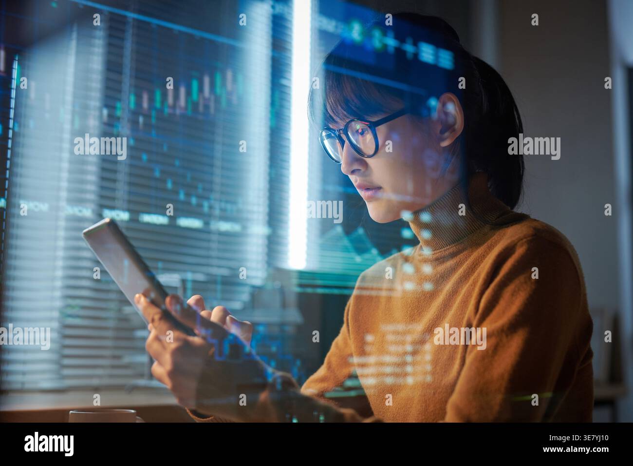Focused Asian business woman wearing eyeglasses using tablet projecting the program sitting at table in office at night Stock Photo