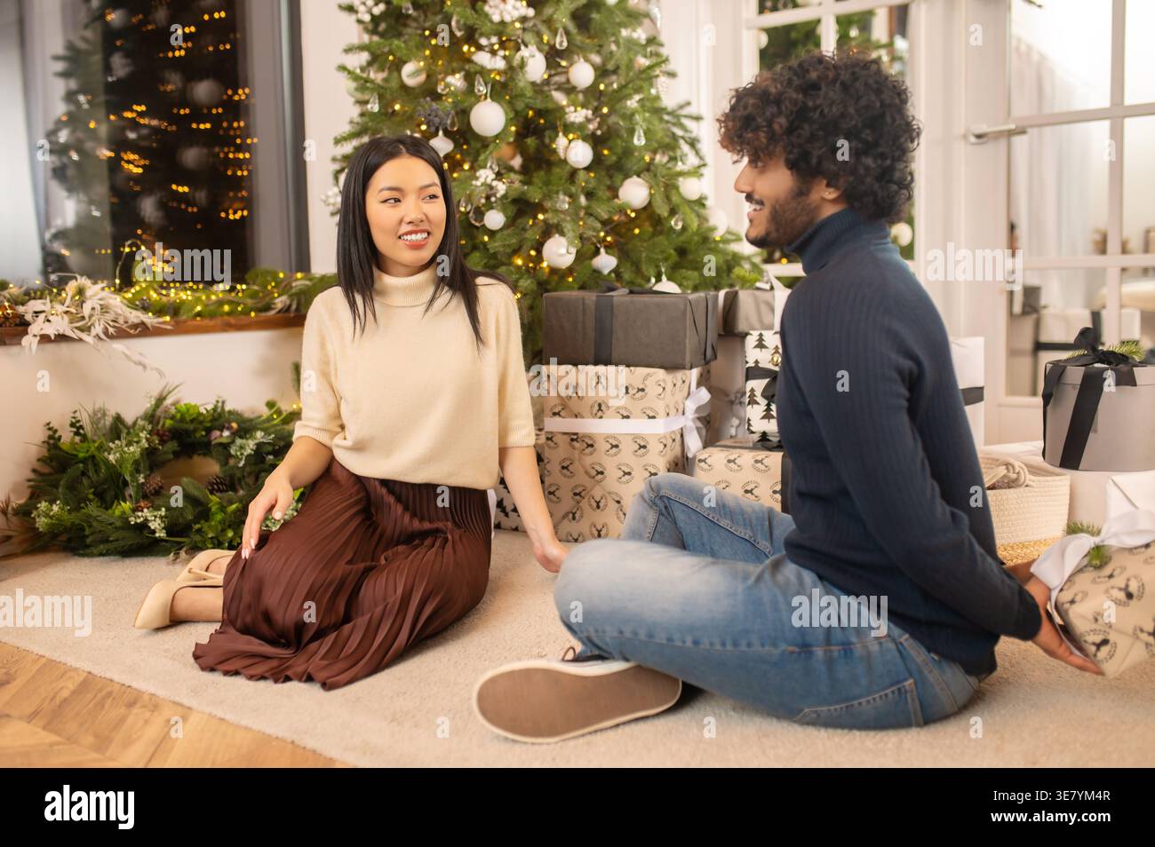 Asian man hiding gift behind sitting with girlfriend on floor during Christmas Stock Photo