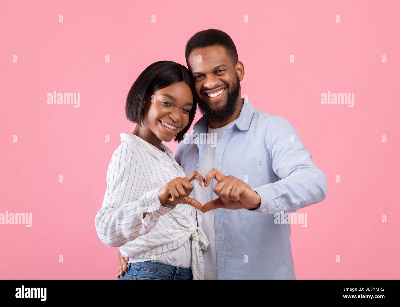 Smiling Black couple making heart with hands while standing against pink background in studio Stock Photo