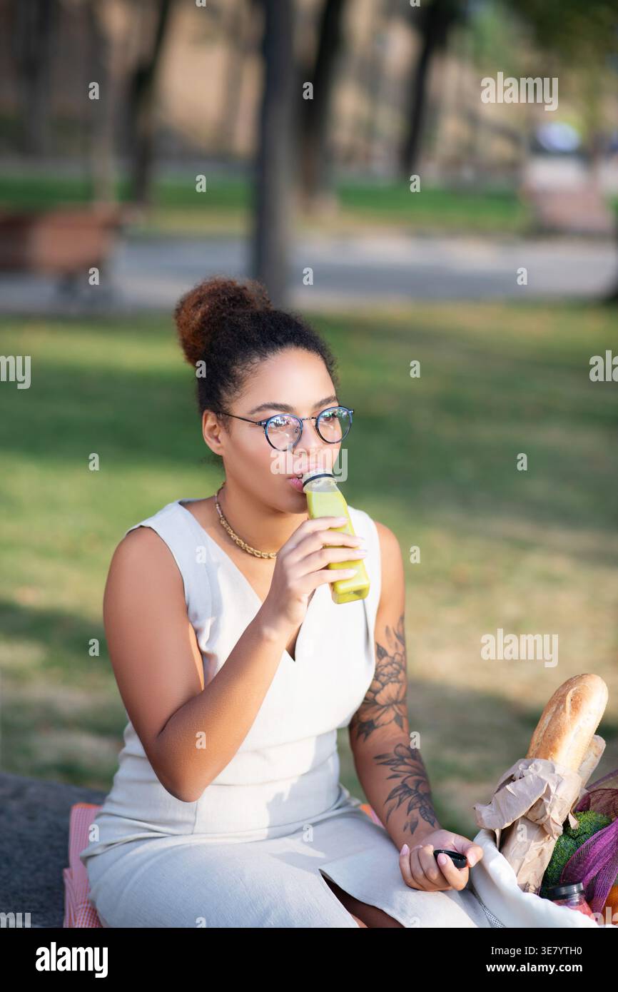Serious Mixed race woman in eyeglasses drinking healthy smoothie looking away sitting on bench near bag with groceries in park on sunny day Stock Photo