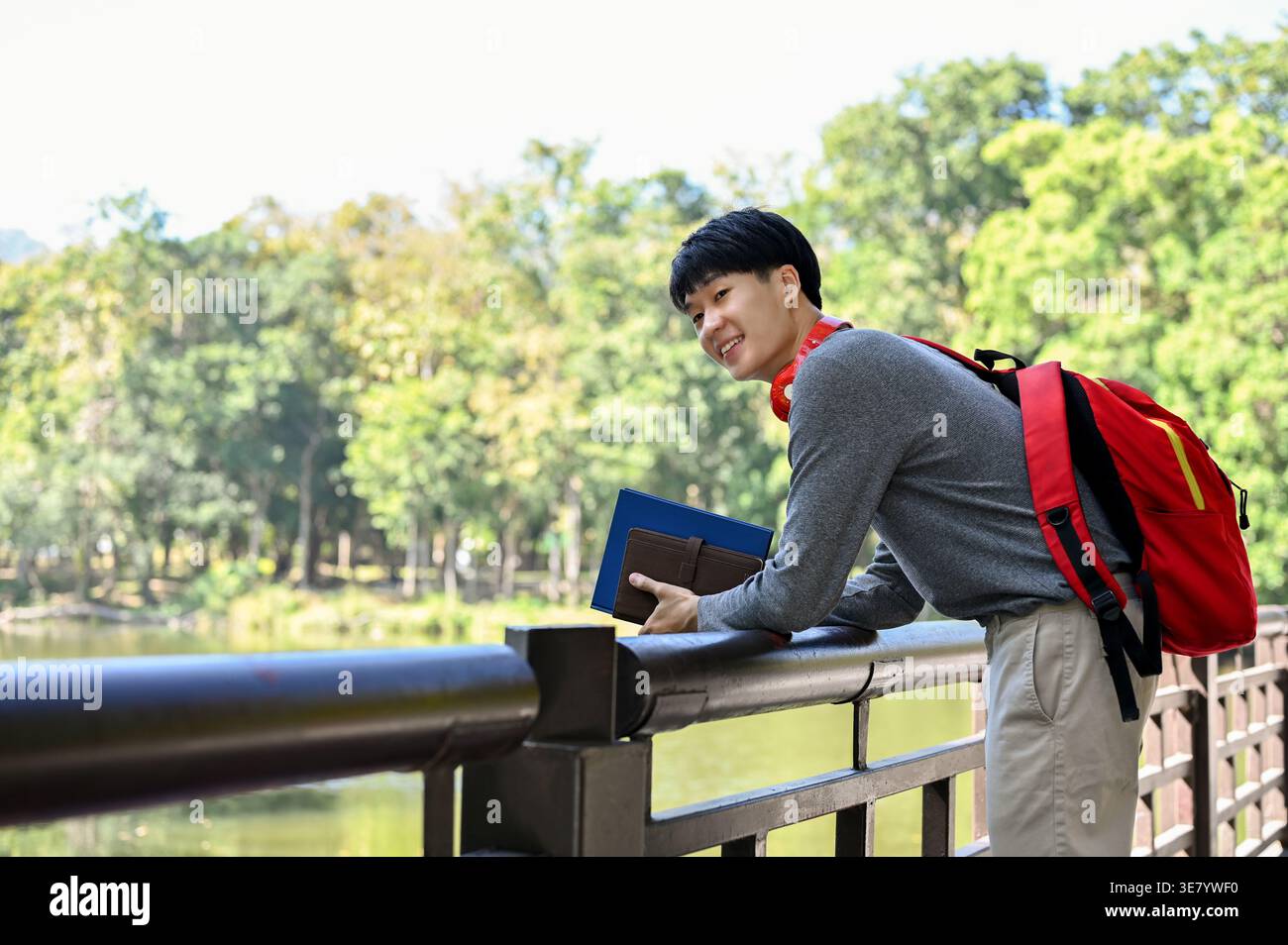 Asian male student holding books looking away while standing with backpack and leaning on metal railing in park during daytime Stock Photo