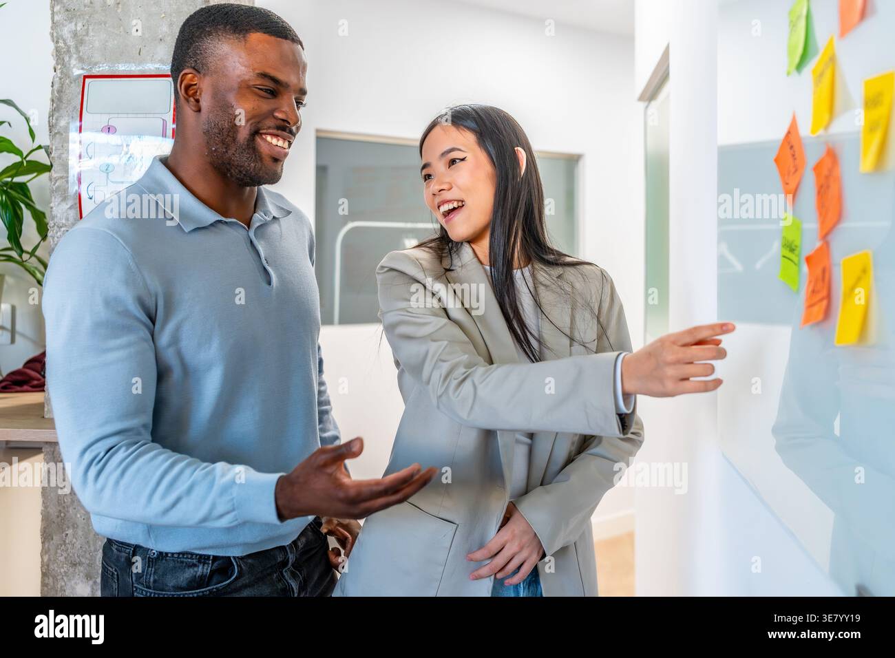 Black male and Asian female colleagues discussing business strategy on whiteboard using sticky notes while standing together in office Stock Photo