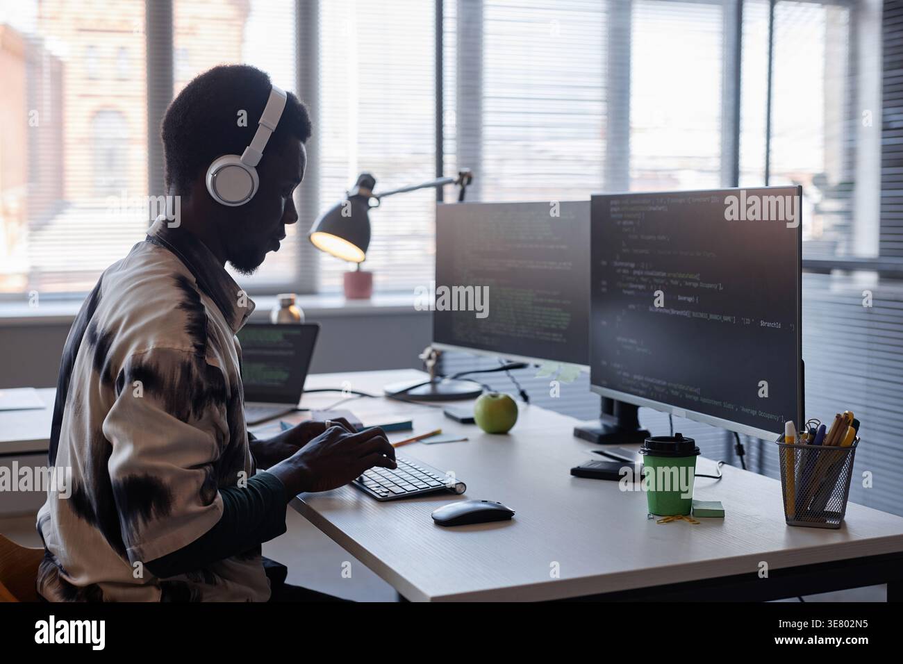 Black male programmer wearing headphones typing codes on computer while sitting at table in office Stock Photo