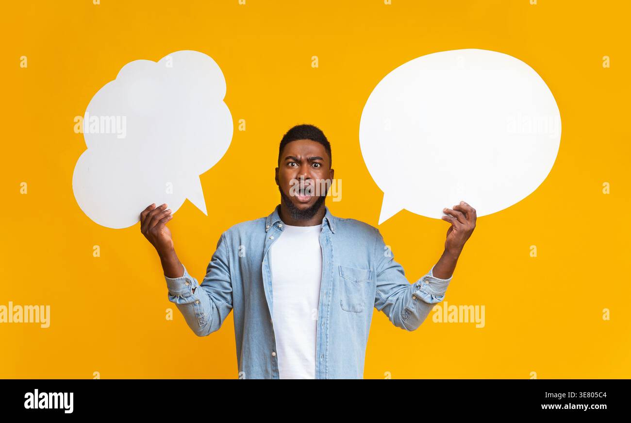 Confused Black man holding two speech bubbles standing against yellow background in studio Stock Photo